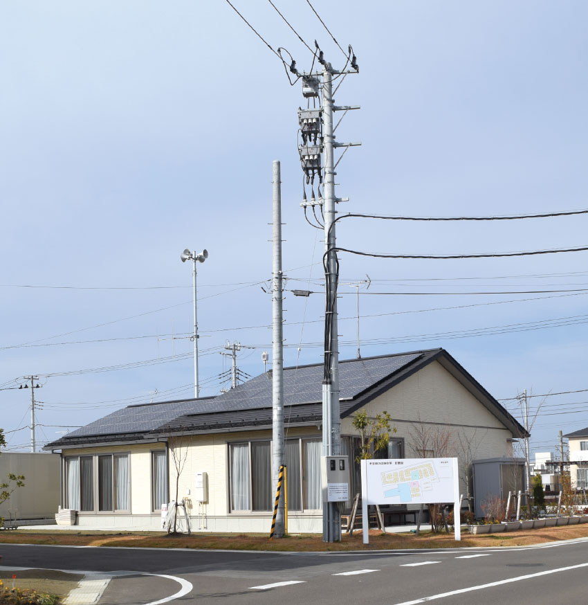 写真9　防災エコタウン内の集会所近くに建てられた第一電柱（低い電柱）。この地点が東北電力からの送電線の最終地点（高い電柱）。電柱の下には東北電力側の2つの高精度電力計（買電用と売電用）が設置されている。